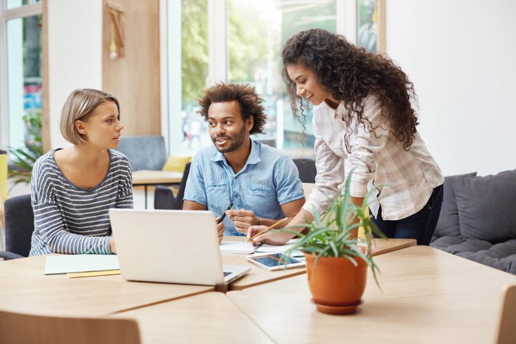 Three young prospective entrepreneurs sitting at library, discussing business plans and company's profits, making business research with laptop, looking through information on tablet